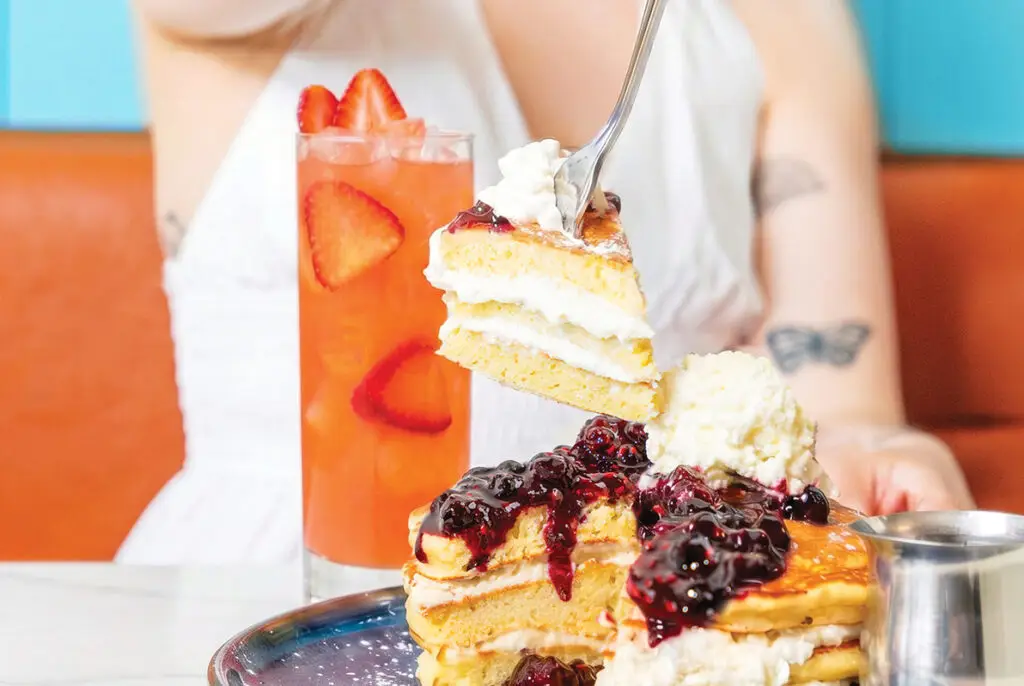 A stack of pancakes with berry compote and whipped cream beside a strawberry drink at OEB Breakfast Co.
